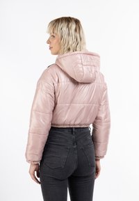 Giacca a vento cropped rosa chiaro con cappuccio, dal finish lucido, polsini elasticizzati e vita aderente. Indossata sopra jeans neri a vita alta.