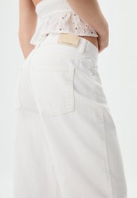 Pantaloni in denim bianchi a vita alta con vestibilità rilassata. Presentano tasche posteriori discrete e una patch con logo sulla cintura. Texture liscia.