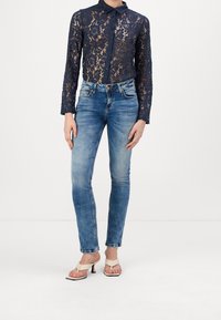 Blouse en dentelle bleu marine à manches longues avec col, fermeture par boutons, associée à un jean skinny bleu clair et des sandales blanches à talons.