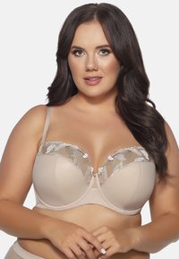 Reggiseno beige con una base liscia e imbottita e un sovraccoperta in pizzo trasparente, caratterizzato da ricami floreali e una chiusura decorativa centrale. Spalline regolabili.