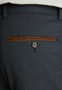 Pantalons bleu marine en tissu texturé, avec une bordure marron au-dessus d'une poche arrière et un bouton gris à quatre trous.