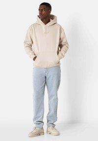 Felpa beige con cappuccio e tasca anteriore a marsupio, abbinata a jeans azzurri chiari e scarpe beige con lacci. Il modello sta in piedi con le mani nelle tasche.