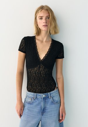 Lyshåret kvinde iført en sort blondebluse med korte ærmer og dyb V-hals samt lyseblå højtaljede jeans, stående op ad en hvid væg.