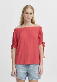 Top rosso off-shoulder con maniche corte e dettagli a fiocco, realizzato in tessuto leggero con una texture arricciata, abbinato a pantaloni corti in denim.