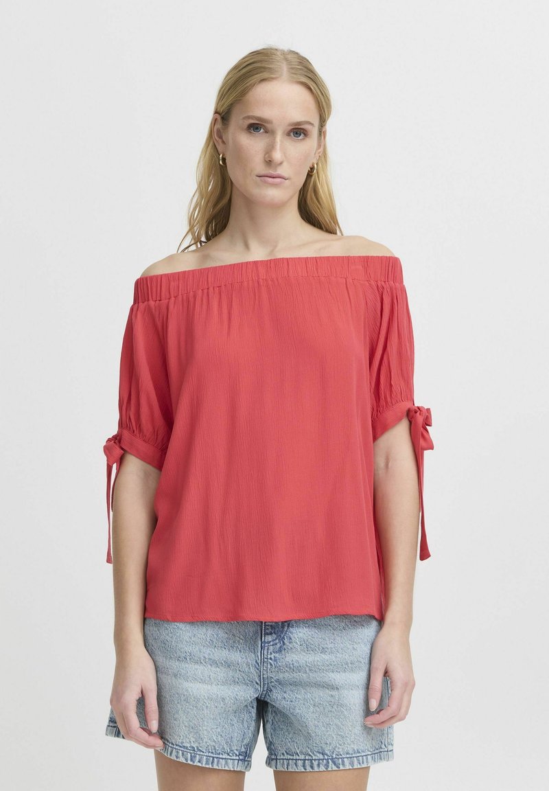 Top rosso off-shoulder con maniche corte e dettagli a fiocco, realizzato in tessuto leggero con una texture arricciata, abbinato a pantaloni corti in denim.