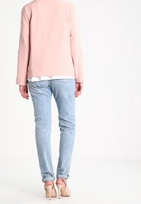 Woman wearing light blue jeans, rolled at cuffs, pale pink blazer, and nude high-heeled shoes, standing facing away against white background.