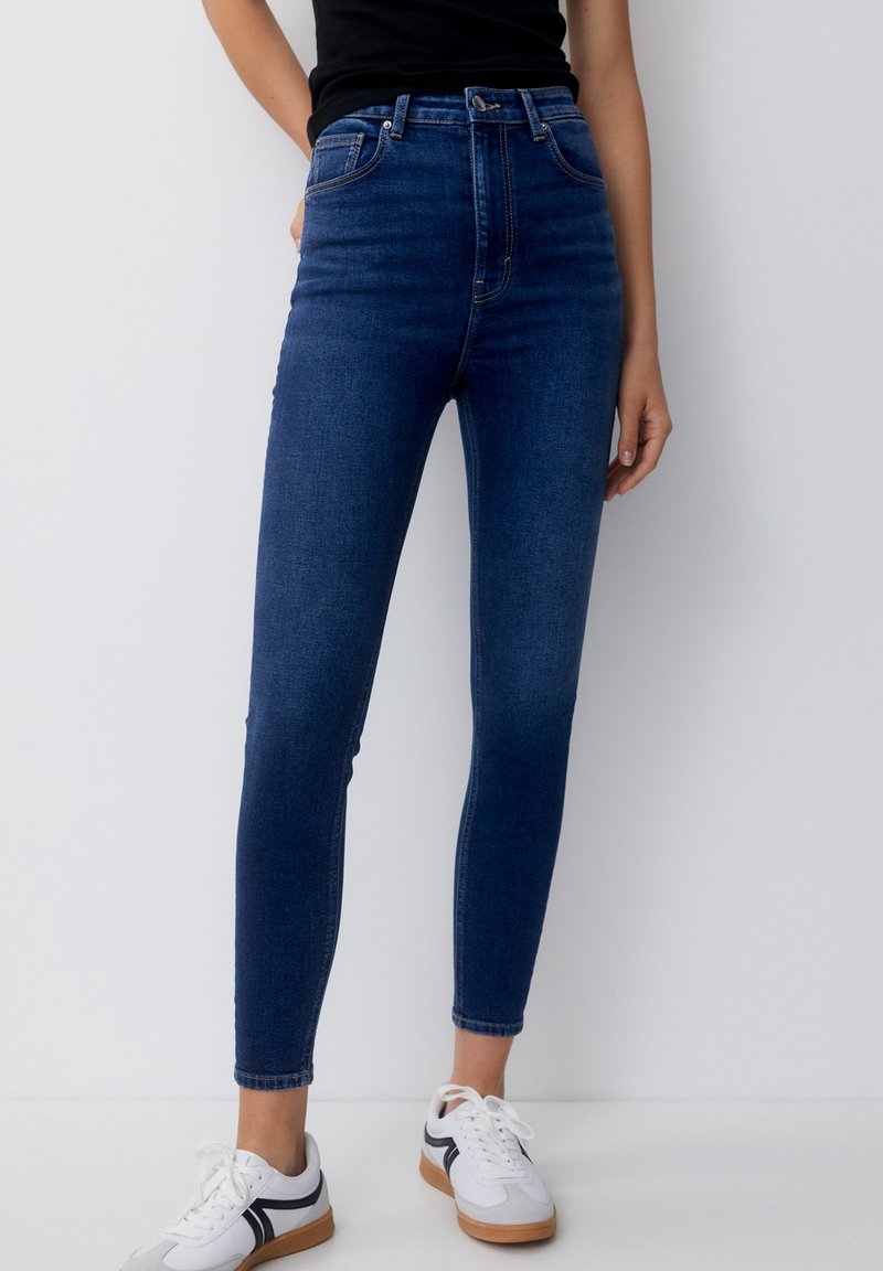 Femme portant un jean skinny taille haute bleu foncé et des baskets blanches avec des bandes noires, debout devant un fond clair uni.