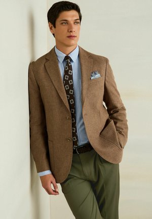 Férfi barna blézert, kék csíkos inget, mintás nyakkendőt és zöld nadrágot visel, világos falnak dőlve.