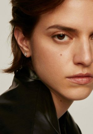 Primer plano de una persona con el cabello corto que lleva una chaqueta de cuero negra. Se puede ver un pequeño pendiente de aro en la oreja derecha. La piel tiene una textura suave.