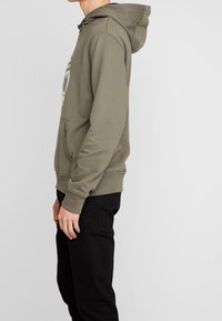 Vista laterale di una persona che indossa un hoodie verde oliva con tasca frontale e pantaloni neri contro uno sfondo chiaro uniforme.