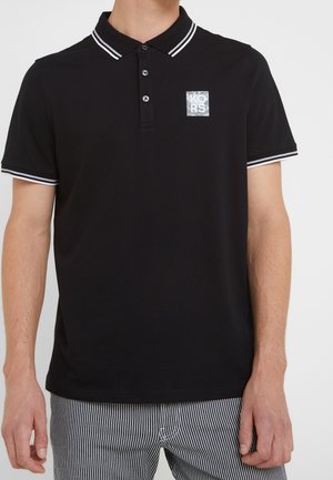 Mężczyzna w czarnej koszulce polo z białymi podwójnymi paskami na kołnierzyku i rękawach, małym kwadratowym logo na lewej piersi oraz w czarno-białe paski spodniach.