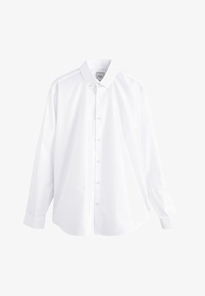 Uśmiechnięty mężczyzna, ubrany w białą koszulę na guziki, ciemne spodnie i czarne eleganckie buty, stoi z jedną ręką w kieszeni na szarym tle.