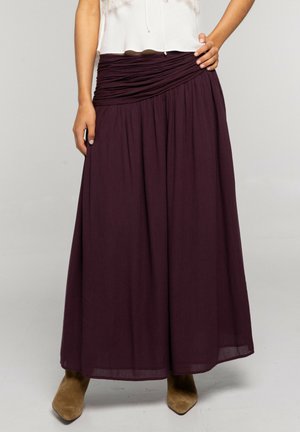 Mujer vistiendo una falda larga plisada de color púrpura oscuro con una cintura fruncida y botas de punta fina color camel visibles debajo del dobladillo.