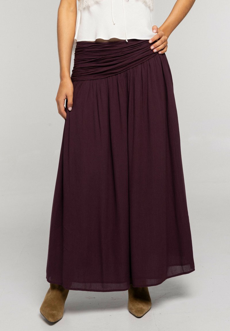 Mujer vistiendo una falda larga plisada de color púrpura oscuro con una cintura fruncida y botas de punta fina color camel visibles debajo del dobladillo.