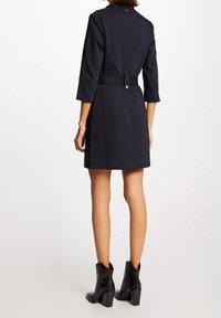 Marinfärgad klänning med figursydd midja, trekvartsärmar och bälte bak. Slät tyg med en strukturerad form. Bärs med svarta ankelskor.