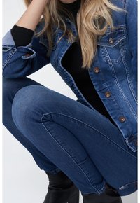 Denim jacket in dark blue with a classic collar, chest pockets, and button closure; paired with dark wash fitted jeans.