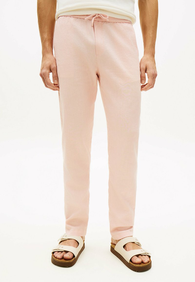 Homme portant un pantalon à cordon de serrage rose clair et des sandales beiges, debout contre un fond blanc.