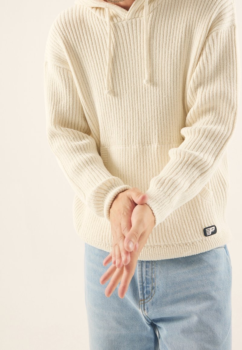 Off-White, gerippter Strickhoodie mit einer Kängurutasche, Kapuze mit Kordelzug und Logo-Patch an der unteren Seite, kombiniert mit hellblauen Jeans.