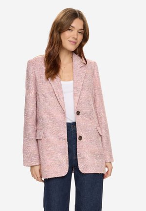 Frau mit langen braunen Haaren, die einen strukturierten rosa Blazer über einem weißen Top und dunkelblauen Jeans trägt, vor einem schlichten Hintergrund stehend.