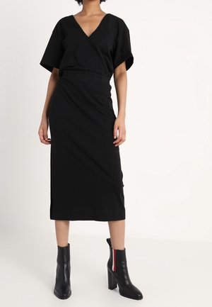Femme portant une robe midi noire à col en V, manches courtes, et des bottines noires avec des bandes latérales rouges et blanches.