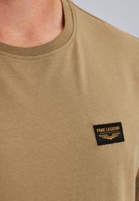 Khaki tričko s kulatým výstřihem. Má černou a žlutou nášivku se štítkem "PME LEGEND". Měkký bavlněný materiál s hladkou texturou.