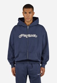 Marineblå zip-up hættejakke lavet af blødt stof, med en stor frontlomme og "PEGADOR" logo i hvid skrift henover brystet.