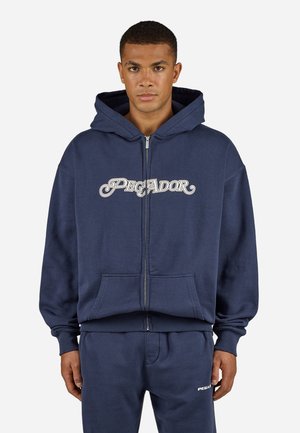 Navy zip-up hoodie made from soft fabric, featuring a large front pocket and "PEGADOR" logo in white script across the chest.