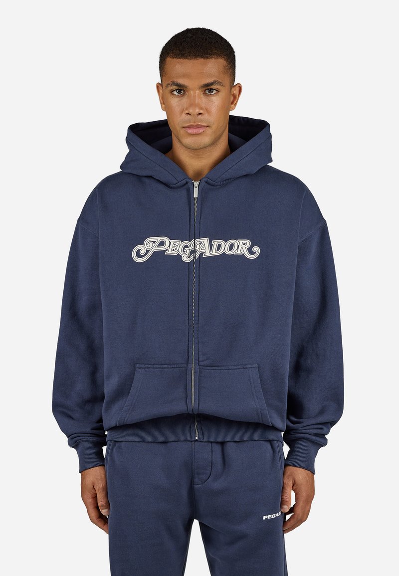 Corta-vento azul-marinho com fecho éclair feito de tecido macio, apresentando um grande bolso frontal e o logótipo "PEGADOR" em script branco no peito.