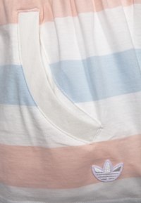 Randig plagg med horisontella band av ljusrosa, vitt och blått. Har en ficka och en liten broderad logotyp i silver längst ner till vänster.