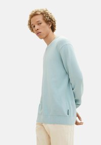 Maglione a maglia testurizzata di un azzurro chiaro, con scollatura rotonda e maniche lunghe. Presenta un orlo a coste e dettagli laterali. Indossato con pantaloni chiari.