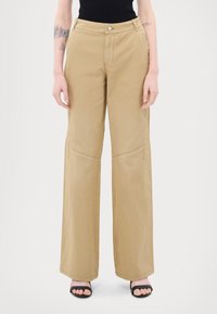 Beige wijde broek van stevig materiaal, met frontzakken, een ritssluiting en stikdetails langs de benen.