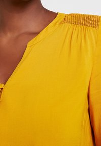 Blusa gialla con scollo a V, dettaglio arricciato testurizzato sulle spalle e tessuto morbido con leggero lucido.