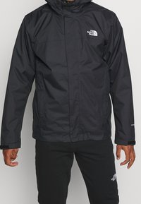 Czarna wodoodporna kurtka z kapturem, zapinana na zamek, z regulowanymi mankietami i logo na lewej piersi. Gładki, lekki materiał.