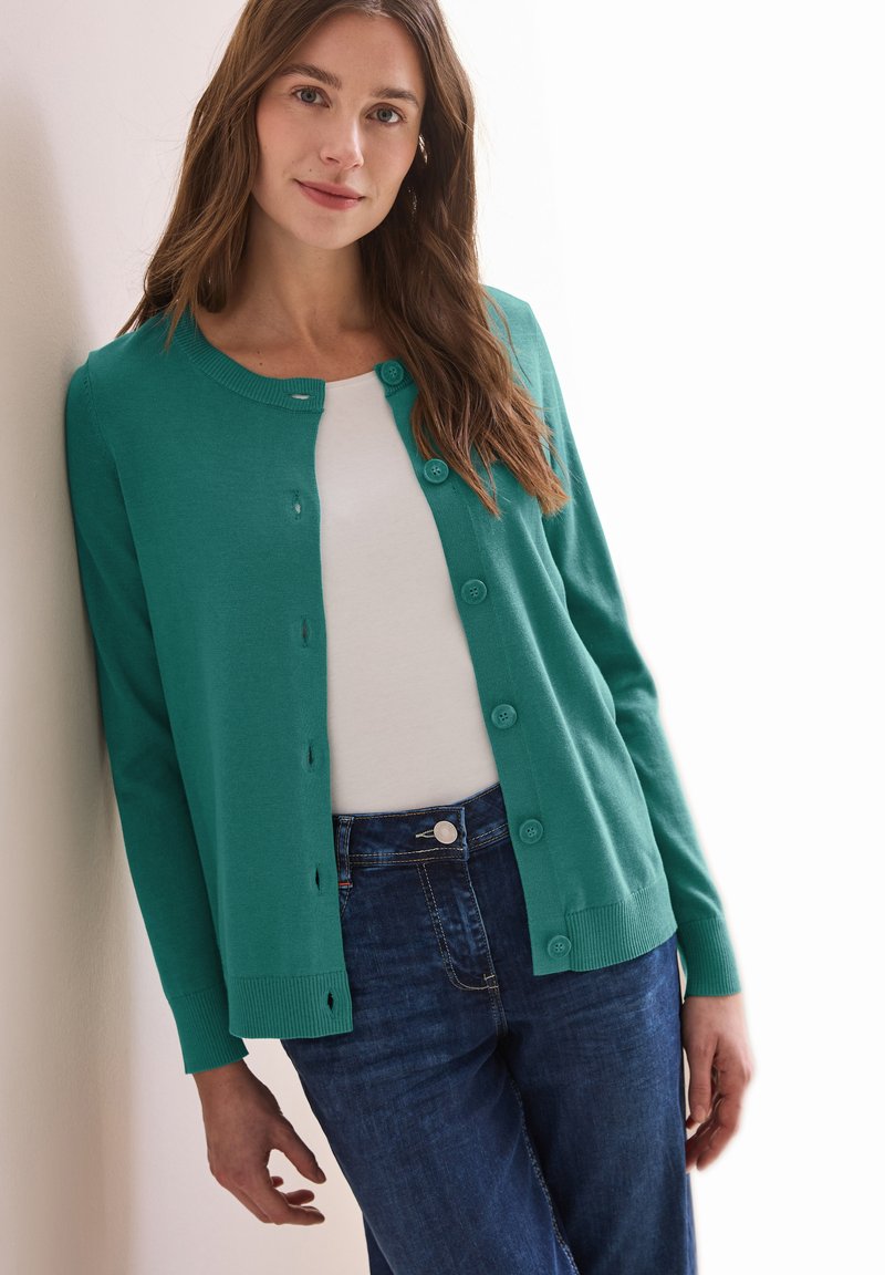 Türkiser Knopfleisten-Cardigan mit Rundhalsausschnitt, gestrickter Textur und gerippten Bündchen, getragen über einem weißen Top mit dunkelblauen Jeans.