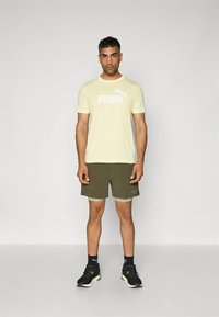 Roupa desportiva masculina: camiseta amarelo pálido com logótipo branco, calções verdes-oliva, ténis pretos com detalhes verdes e meias pretas até ao tornozelo.