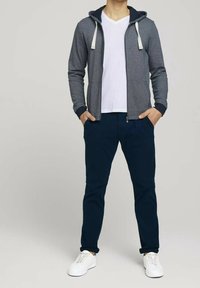 Felpa grigia con zip e lacci bianchi, indossata sopra una maglietta bianca, abbinata a pantaloni blu navy e sneakers bianche. Tessuto uniforme.