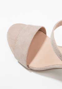 Beżowe zamszowe sandały z szerokim paskiem na palce i wąskim paskiem na kostkę, z gładką podeszwą i stonowanym wzornictwem.