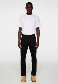 Črne kavbojke s pravim krojem, kombinirane z preprosto belo majico. Model nosi svetlo rjave škornje in stoji pred enobarvnim ozadjem.