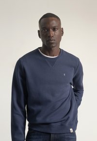 Sudadera azul marino con cuello y puños acanalados, hecha de material suave. Cuenta con un pequeño logo en el pecho y una etiqueta en el dobladillo.