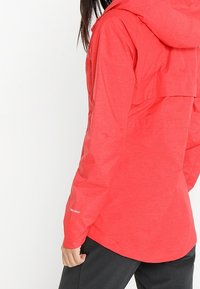 Veste à capuche rouge avec des manches longues, une coupe décontractée et un détail de rabat à l'arrière. Le tissu semble léger et légèrement texturé.