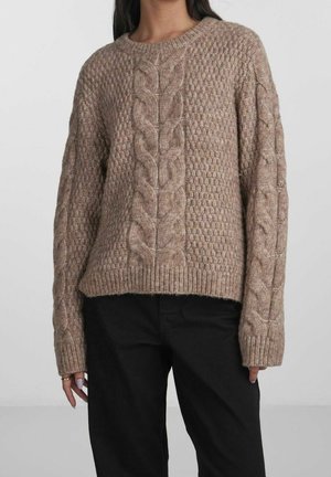 Femme portant un pull en maille épaisse beige avec des motifs torsadés sur le devant et les manches, associé à un pantalon noir, debout devant un fond clair.