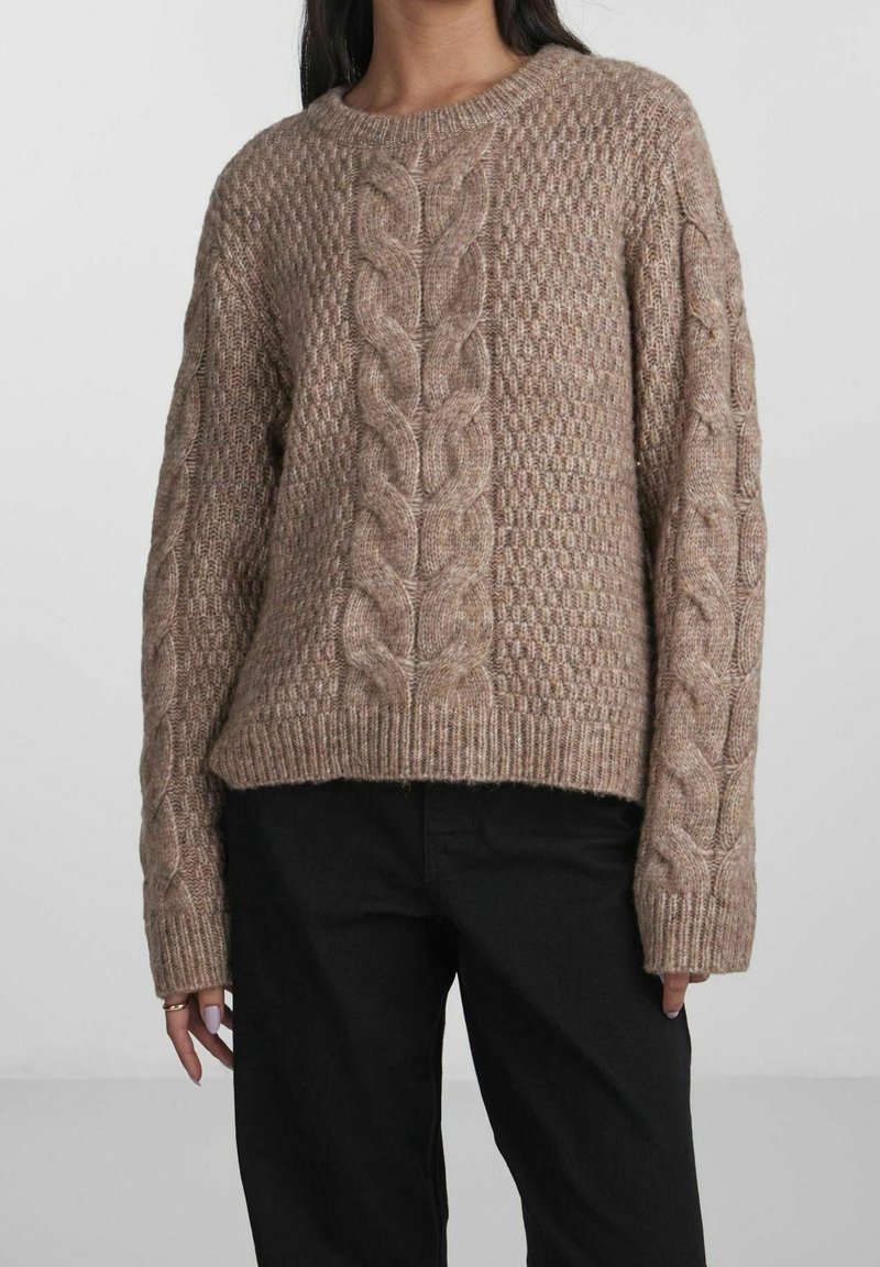 Femme portant un pull en maille épaisse beige avec des motifs torsadés sur le devant et les manches, associé à un pantalon noir, debout devant un fond clair.