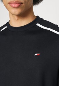 T-shirt nera a maniche corte con collo rotondo, caratterizzata da accenti bianchi sulle spalle e un piccolo logo in rosso, bianco e blu.