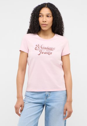 Woman with curly hair wearing a light pink Mustang Jeans t-shirt and light blue jeans, looking slightly to the side.