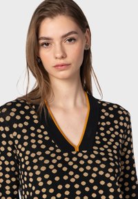 Maglione nero con pois beige, scollo a V e rifiniture arancioni a contrasto. Texture morbida, design aderente e dettagli sottili in tutto il capo.