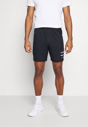 Homme portant un short noir Tommy Sport, un t-shirt blanc, des chaussettes blanches et des baskets blanches, debout devant un fond clair uni.