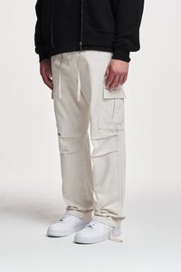 Pantaloni cargo beige con molte tasche laterali, vita regolabile con cordino e orli a coste, abbinati a sneakers bianche su uno sfondo semplice.