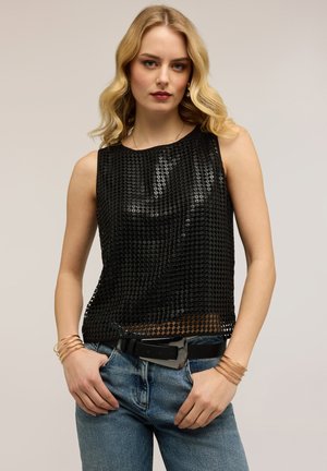 Blonde Frau mit welligem Haar trägt ein schwarzes ärmelloses Paillettenoberteil, blaue Jeans mit schwarzem Gürtel und goldene Armreifen, steht vor neutralem Hintergrund.