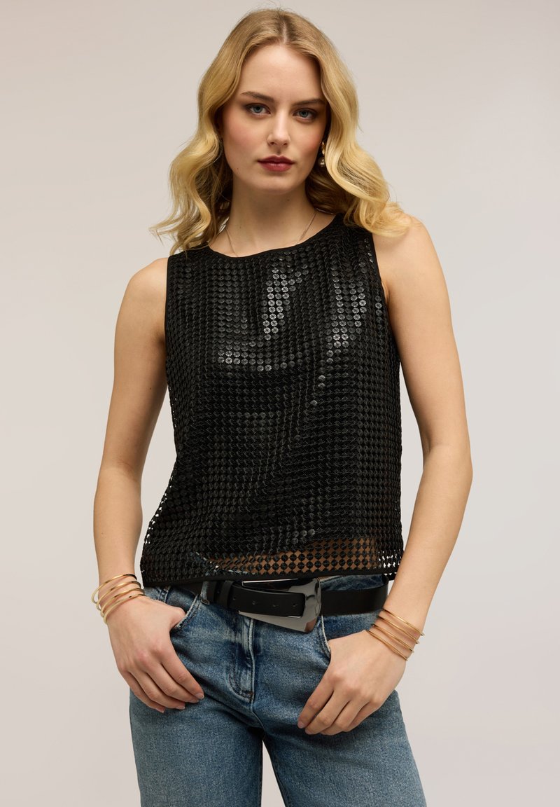 Mujer rubia con pelo ondulado lleva una blusa sin mangas de lentejuelas negra, jeans azules con cinturón negro y pulseras doradas, de pie contra un fondo neutro.