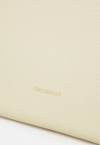 Cremefärgad läderhandväska med pebblemönster. Utrustad med präglad guld "Coccinelle" logotyp. Fokuserar på material och subtila designdetaljer.